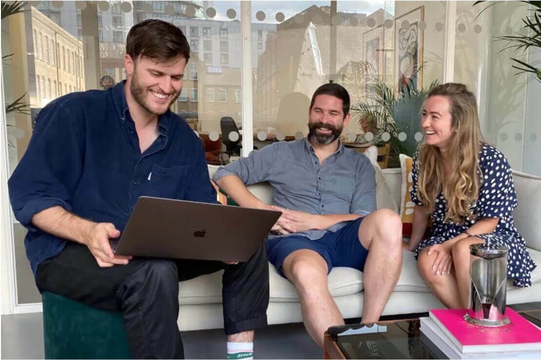 A team of three people sat on a sofa smiling whilst looking at a laptop